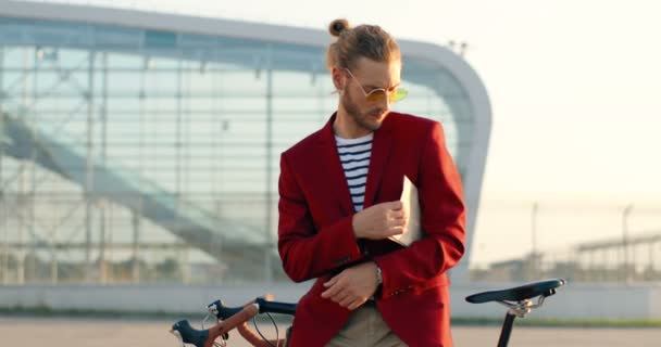 Élégant jeune homme caucasien en veste rouge et lunettes de soleil assis sur le vélo et la lecture de journaux en plein air. Homme dans un style décontracté appuyé sur le vélo et lire le papier. À l'aéroport à l'extérieur. Paysage urbain. — Video