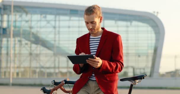 Caucasien jeune homme sourire beau debout à vélo et en utilisant un appareil tablette. Élégant gars en veste rouge appuyé sur le vélo et tapoter ou défiler sur l'ordinateur gadget au paysage de la ville à l'aéroport. — Video