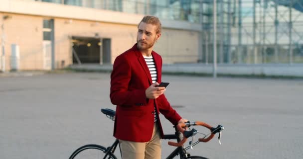 Blanke mannelijke fietser in casual stijl en rode jas leidende fiets en tikken of scrollen op smartphone op straat. Knappe jongeman die met de fiets loopt en sms 't op de mobiele telefoon. — Stockvideo