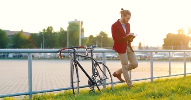 Blanke stijlvolle mannelijke fietser staan op straat, leunen en schrijven plannen in notebook. Knappe man in casual stijl noteren in planner en denken outdoor op de fiets. — Stockvideo