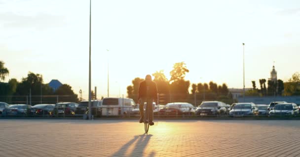 Caucasico sorrise gioioso ciclista maschile al tramonto all'aperto. Elegante bell'uomo in sella alla bicicletta e attraversando parcheggio auto. Auto sullo sfondo. Ragazzo allegro che fa un giro in bella serata estiva in città. — Video Stock