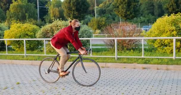 Caucasico giovane elegante ciclista maschile in giacca casual rossa e maschera in sella a una bicicletta in strada in città. Concetto di pandemia di coronavirus. Bell'uomo in protezione respiratoria con giro in bicicletta. — Video Stock