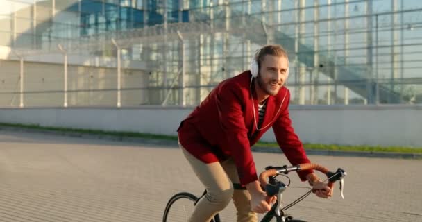 Blanke vrolijke jonge stijlvolle mannelijke fietser in rood casual jasje en koptelefoon rijden op een fiets op straat. Knappe vrolijke glimlachende man die fietstocht maakte en naar muziek luisterde. Stadslandschap — Stockvideo