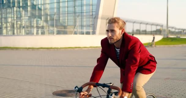 Blanke jonge stijlvolle mannelijke fietser in rood casual jasje rijden op een fiets op straat in de stad. Knappe man met een fietstocht. Glas modern gebouw op achtergrond. Stedelijk landschap. — Stockvideo