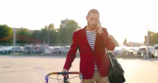 Blanke mannelijke fietser in casual stijl en rode jas holding bike en praten op mobiele telefoon op straat. Knappe jongeman met fiets en spreken op mobiele telefoon. Auto 's parkeren op achtergrond. — Stockvideo
