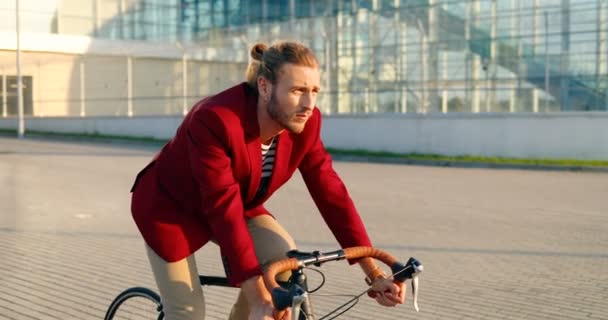 Blanke stijlvolle mannelijke fietser in rood casual jasje rijden op een fiets op straat in de stad. Knappe man met oortjes die fietsen. Glas modern gebouw op achtergrond. Stedelijk landschap. Zonnige dag. — Stockvideo