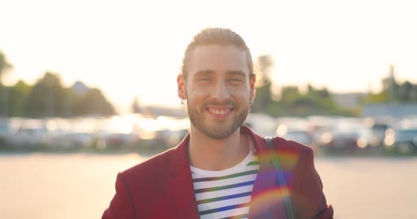 Portrait de jeune homme blanc gai beau en veste décontractée rouge souriant à la caméra à l'extérieur au parking. Gros plan de joyeux souriant homme heureux sur une journée ensoleillée. Voitures en arrière-plan. — Video