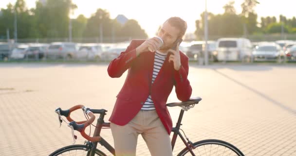 Man casual knappe fietser in rode jas leunend op de fiets, nippen warm drankje en praten op mobiele telefoon. Bij groot glas modern gebouw. Blanke man die koffie drinkt en spreekt op mobiele telefoon. — Stockvideo