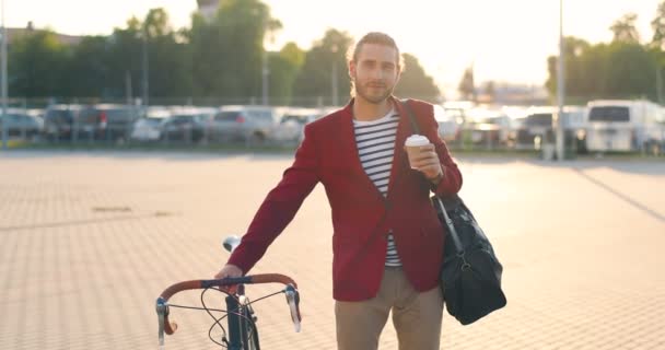 自転車にもたれて、ホットドリンクを保持し、カメラに笑みを浮かべて赤いジャケットの男性のカジュアルなハンサムな自転車のライダーの肖像画。駐車場で。コーヒーとコーヒー男とともに車の背景. — ストック動画