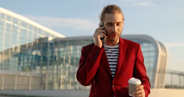 Casual hombre guapo feliz en chaqueta roja de pie al aire libre, bebiendo bebida caliente y hablando por teléfono celular. En el gran edificio de cristal moderno. Hombre caucásico bebiendo café, sonriendo y hablando por teléfono móvil — Vídeos de Stock