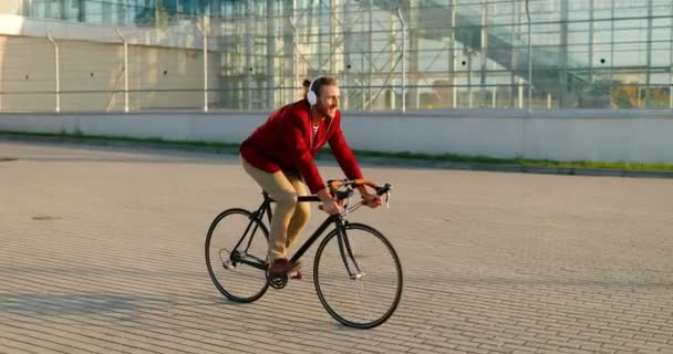 Kaukasischer junger, stylischer männlicher Radfahrer in roter Freizeitjacke und Kopfhörer, der auf der Straße in der Stadt Fahrrad fährt. Schöner Mann beim Fahrradfahren und Musikhören. Stadtlandschaft. Draußen. — Stockvideo