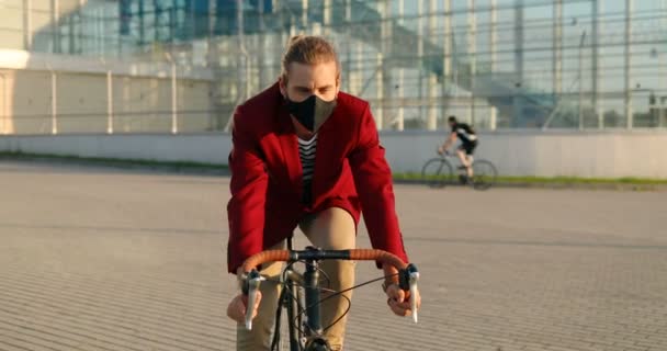 Bicicleta masculina de estilo caucásico con chaqueta casual roja y máscara que monta una bicicleta en la ciudad. Hombre guapo en protección respiratoria teniendo paseo en bicicleta. Edificio moderno de vidrio sobre fondo. Paisaje urbano. — Vídeos de Stock