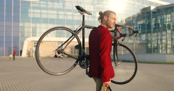 Caucásico guapo joven elegante hombre en chaqueta casual roja caminando al aire libre y llevando bicicleta en sholder. Paisaje urbano. Edificio de vidrio moderno sobre fondo. Jinete masculino paseando y sosteniendo la bicicleta. — Vídeos de Stock