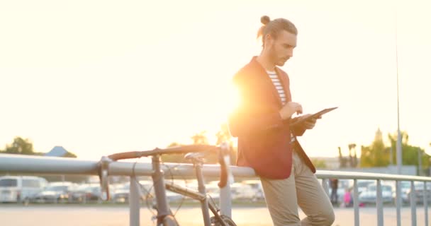 Νεαρός καυκάσιος όμορφος άνδρας με κόκκινο σακάκι στέκεται έξω στο ηλιοβασίλεμα σε χώρο στάθμευσης αυτοκινήτων με ποδήλατο και χρησιμοποιώντας συσκευή tablet. Αρσενικός ποδηλάτης σκέψης και χτυπώντας, κύλιση ή γραπτών μηνυμάτων στον υπολογιστή gadget — Αρχείο Βίντεο
