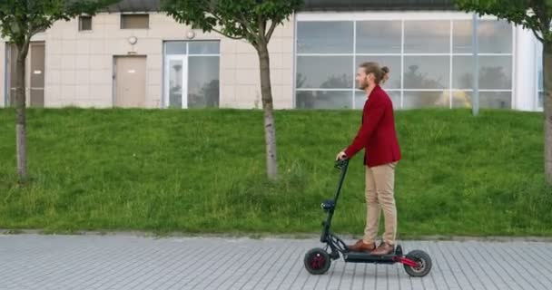 Vit glad snygg ung man i röd jacka rider elektrisk skoter och står stilla på gatan i staden. Snygg manlig ryttare ler och har roligt medan du går någonstans. Stadsresa. — Stockvideo