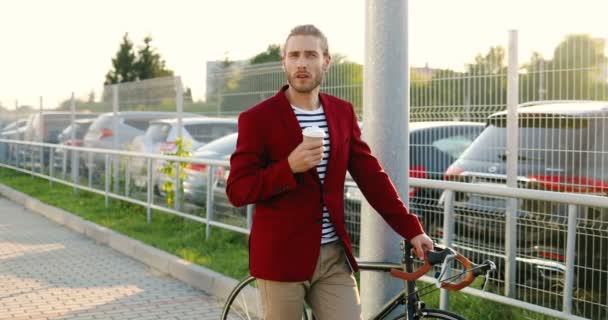 Caucasico bello giovane uomo alla moda in piedi al parcheggio auto con bicicletta, riposo e sorseggiando bevanda calda. Attraente sosta ciclista maschile e bere caffè all'aperto. — Video Stock