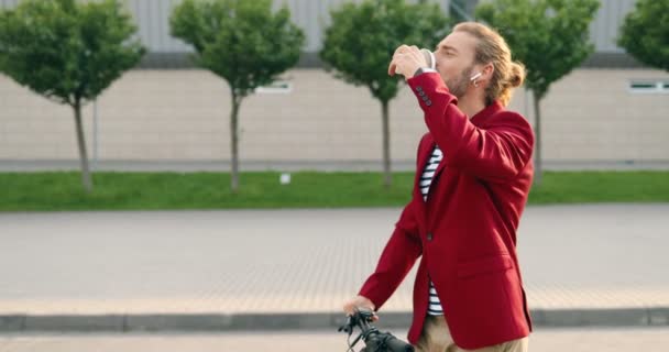 Przystojny biały, młody, stylowy mężczyzna w słuchawkach i czerwonej kurtce jeżdżący na skuterze elektrycznym na miejskiej ulicy i pijący kawę. mężczyzna jeździec stojąc, sącząc drinka i idąc w miasto na drodze. — Wideo stockowe