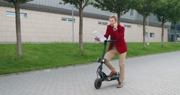 Kaukázusi, fiatal, elegáns üzletember áll az elektromos robogónál, mobilon beszél és dokumentumokat tart magánál. A szabadban. Jóképű férfi beszél mobilon és szerződést olvas. Üzleti megbeszélések. — Stock videók