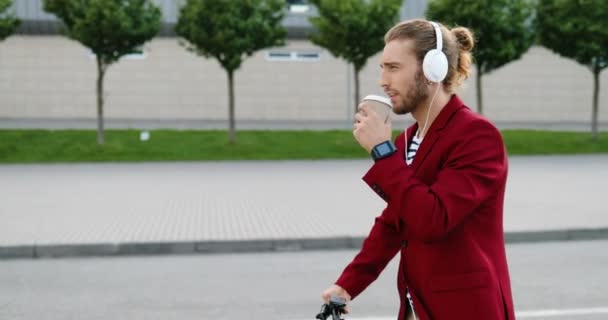 Caucásico joven y elegante hombre guapo en los auriculares que conducen scooter eléctrico en la calle, escuchando música y bebiendo. Un tipo montando un vehículo y tomando café al aire libre. En paseo por la ciudad. — Vídeo de stock