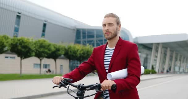 Ritratto di giovane caucasico bello in giacca rossa in piedi all'aperto con bici o scooter elettrico e guardando la fotocamera. Bello ragazzo alla moda in bicicletta in possesso di carta o disegno sulla strada della città — Video Stock