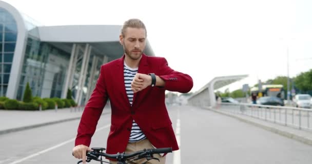 Кавказький молодий стильний чоловік у червоній куртці їде на електричному скутері і шукає час за ручним годинником. На вулиці. Поганий водій їде і кидається в місто. Попередня концепція. — стокове відео