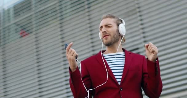 Kavkazský mladý stylový pohledný muž ve sluchátkách, který poslouchá hudbu na chytrém telefonu a zpívá. Venkovní. Veselý, dobře vypadající vtipálek v červeném saku zpívá a poslouchá písničku. Hráč mobilního telefonu. — Stock video