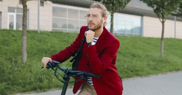 Retrato de joven guapo caucásico en chaqueta roja y gafas de pie al aire libre con scooter eléctrico y mirando a la cámara. Un tipo elegante en el vehículo en la calle de la ciudad. Dolly disparó. Acercamiento. — Vídeo de stock