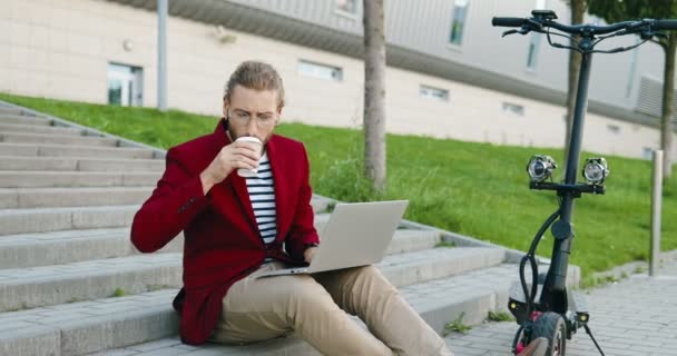 Jovem caucasiano bonito homem de óculos e casaco vermelho sentado em degraus ao ar livre, trabalhando no computador portátil, bebendo. Um tipo elegante a beber café. Freelancer. Scooter elétrico. — Vídeo de Stock