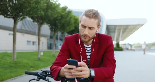 Hvit, ung, kjekk mann i rød jakke som står på gata på elektrisk scooter og hører på musikk på mobilen. Stilig fyr som avlytter smarttelefon med hodetelefoner. SMS på mobiltelefon. – stockvideo