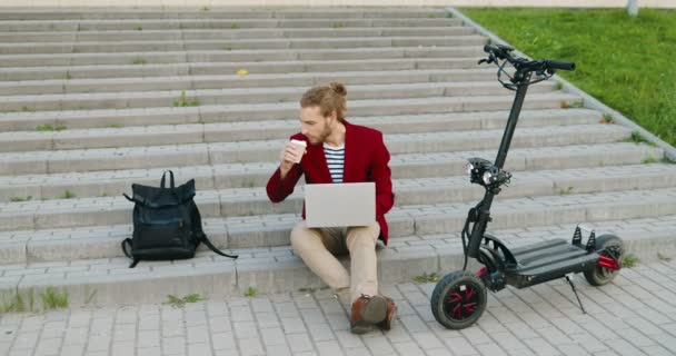穿着红色夹克的年轻的高加索帅哥坐在外面的台阶上，在笔记本电脑上工作，喝着酒。时髦的家伙喝咖啡。忙碌的自由职业者。电动滑板车自由职业. — 图库视频影像