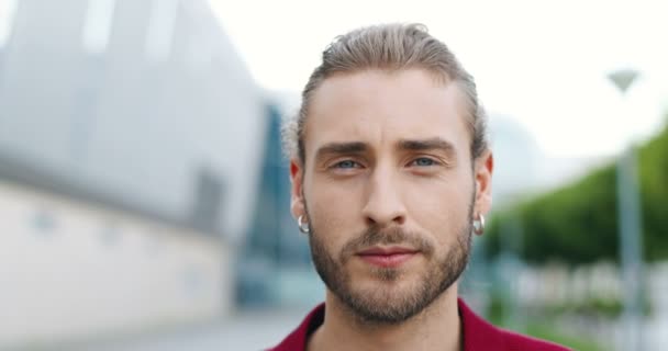 Ritratto di giovane uomo caucasico con la barba che guarda la telecamera all'aperto. Primo piano di faccia di bel maschio elegante con due orecchini in orecchi a strada. — Video Stock