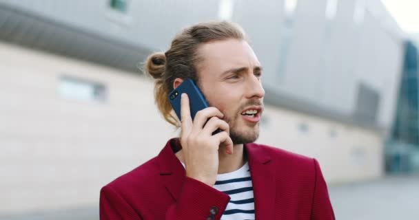 Gros plan de jeune homme blanc beau en veste rouge parlant sur téléphone mobile à l'extérieur. Sérieux beau mâle élégant debout dans la rue et parlant sur téléphone portable. Conversation téléphonique. — Video