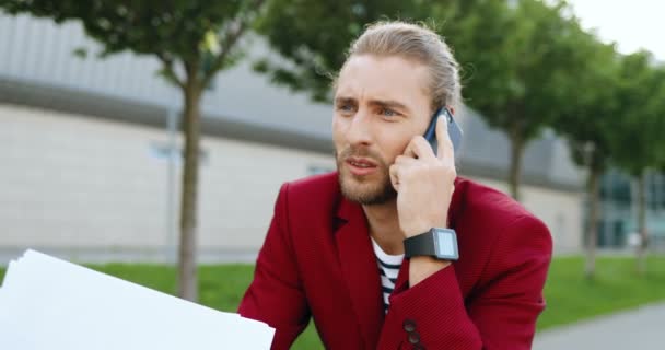 Közelkép kaukázusi fiatalember piros kabátban és okos óra beszél a mobiltelefon szabadtéri és olvasási szerződést. Stílusos üzletember olvasta és nézte dokumentumok, miközben beszélt a mobiltelefon. — Stock videók