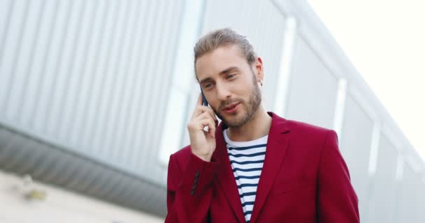 Junger gutaussehender, glücklicher Kaukasier in roter Jacke, der draußen mit dem Handy telefoniert. Fröhlicher, gut aussehender, stylischer Mann, der auf der Straße steht und mit dem Handy spricht. Telefonat. Aufruf. — Stockvideo