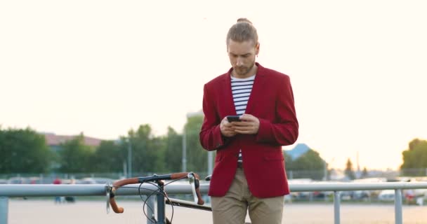 Vit ung snygg stilig man står på gatan med cykel och sms: a meddelande på smartphone. Manlig tappning och rullning på mobiltelefon med cykel utanför. — Stockvideo