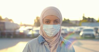 Geleneksel türban ve tıbbi maske takmış, kameraya bakan genç Müslüman güzel bir kadının portresi. Güneş ışığında sokağın ortasında tesettürlü Arap kadın. Arap kız..