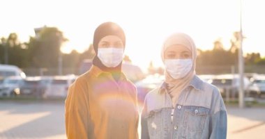 Geleneksel türbanlı ve tıbbi maskeli güzel mutlu Arap kadınlarının güneş ışığına bakan portresi. Açık havada güneşli bir yaz. Müslüman, neşeli, tesettürlü kadınlar. Şehirde İslamcı kızlar.