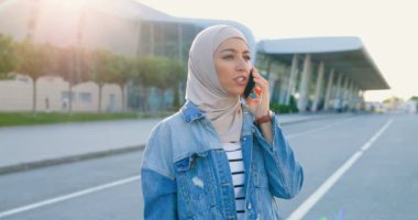 Geleneksel başörtüsü takmış Arap genç ve güzel bir kadın sokakta duruyor ve cep telefonuyla konuşuyor. Müslüman neşeli kız cep telefonundan konuşuyor. Büyük, modern cam binalı şehir manzarası.