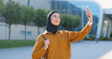 Genç Müslüman, siyah tesettürlü güzel bir kadın caddede dikiliyor ve akıllı telefonuyla selfie çekiyor. Arap güzel bayan gülümsüyor ve cep telefonunun kamerasına poz veriyor. Fotoğrafları selfie.