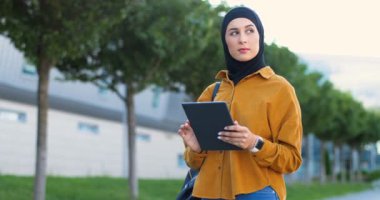 Siyah başörtüsü takmış Arap genç ve güzel bir kadın sokakta duruyor ve tablet kullanıyor. Geleneksel tesettürlü şık Müslüman kadın. Alet bilgisayarında tıkırdıyor ve kaydırıyor. Açık havada..