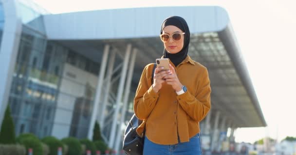 携帯電話をタップし 陽気に笑みを浮かべて通りで黒ヒジャーブとサングラスのイスラム教徒の美しい女性 街の外の携帯電話でアラビア語かなりスタイリッシュな女性のテキストメッセージ メッセージ — ストック動画