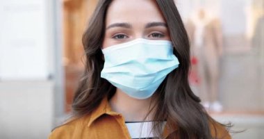 Medikal maskeli güzel bir kadının portresi kameraya bakıyor ve neşeyle gülümsüyor. Coronavirus konsepti. Açık havada. Solunum korumasındaki güzel kızın yüzünü kapatın..