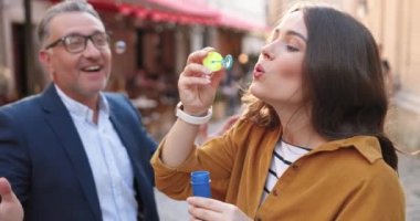 Beyaz, yetişkin, güzel bir kız sokakta para ödüyor ve orta yaşlı bir babayla mutlu bir gün geçirirken sabun köpüğü üflüyor. Açık havada. Babam gözlüklü gülümsüyor ve güzel kızıyla eğleniyor..