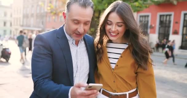 Happy Caucasian father walking with adult beautiful daughter, using smartphone and talking. Having talk. Outdoor. Cheerful man and girl strolling, watching on mobile phone and smiling. Cozy street. — Stock Video