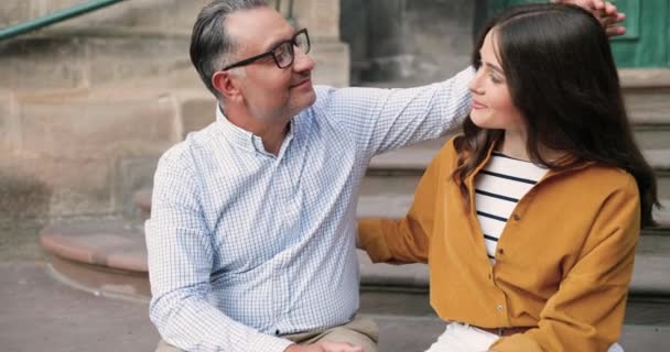 Sonrió feliz padre caucásico en gafas abrazando bonita hija adulta al aire libre mientras se sentaban en los escalones. Hermosa joven alegre sonriendo en abrazos de papá. Amor de la paternidad. — Vídeos de Stock