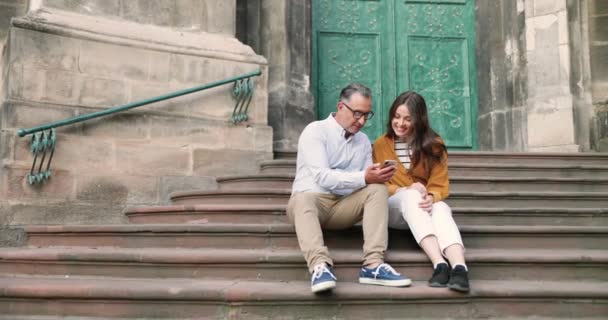 Bastante joven chica caucásica con el padre sentado en los escalones de la iglesia y el uso de teléfono inteligente al aire libre. Hombre de mediana edad mostrando algo en el teléfono móvil a hermosa hija adulta, discutiendo y hablando. — Vídeos de Stock