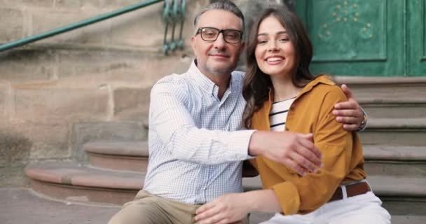 Retrato de feliz padre caucásico en gafas abrazando bonita hija adulta al aire libre mientras se sienta en los pasos amd sonriendo a la cámara. Hermosa joven alegre sonriendo en abrazos de papá. Amor de la paternidad. — Vídeo de stock