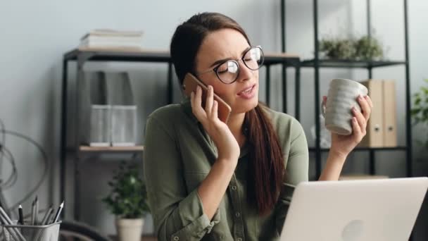 Retrato de joven hermosa mujer con gafas de trabajo en el ordenador portátil. Chica cansada hablando por teléfono y tomando café de la taza mientras está sentada en el escritorio. — Vídeo de stock