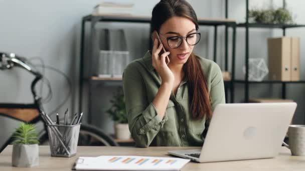 Retrato de una joven hermosa mujer trabajando en un portátil. Chica segura consulta al cliente por teléfono en busca de información sobre el ordenador portátil. — Vídeos de Stock