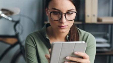 İş yerinde tablet kullanan gözlüklü güzel bir kıza yakın durun. Genç kadın elektronik alet kullanıyor ve gülümsüyor..